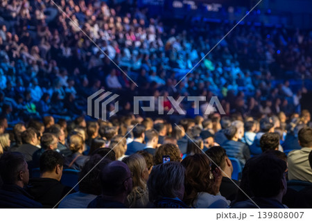 Audience at packed conference venue watching presentation under blue stage lighting Audience at packed conference venue watching presentation under blue stage lighting 139808007