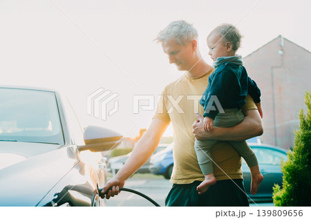 Young man holding baby and install cable plug to his electric car with home charging station. Residential EV Charging Station. Sustainable Power of a Wall-Mounted Charger. Future of Clean Transport. Young man holding baby and install cable plug to his electric car with home charging station. Residential EV Charging Station. Sustainable Power of a Wall-Mounted Charger. Future of Clean Transport. 139809656