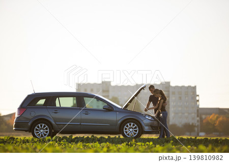 Young couple, handsome man and attractive woman at car with popped hood checking oil level in engine using dipstick on clear sky background. Transportation, vehicles problems and breakdowns concept. Young couple, handsome man and attractive woman at car with popped hood checking oil level in engine using dipstick on clear sky background. Transportation, vehicles problems and breakdowns concept. 139810982