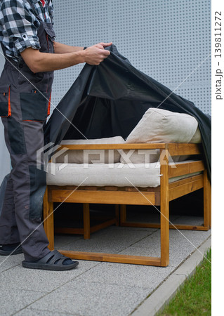 Man removes protective cover from outdoor sofa with cushions on patio. Concept of garden furniture care 139811272