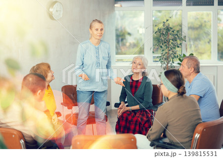 Older woman presides over support group meeting with mature people listening Older woman presides over support group meeting with mature people listening 139815531