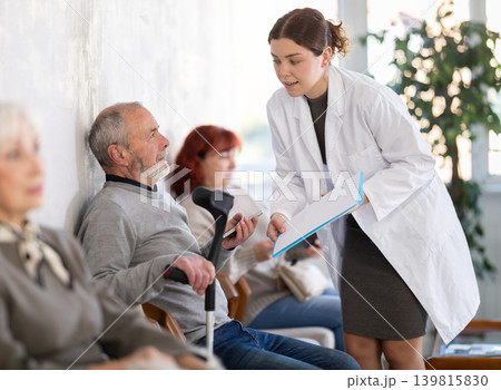 Positive polite nurse or doctor talking to aged man patient in reception area in background queue of people 139815830