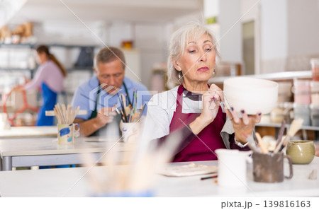 Woman potter with paintbrush, painting on plate in workshop, working in studio Woman potter with paintbrush, painting on plate in workshop, working in studio 139816163