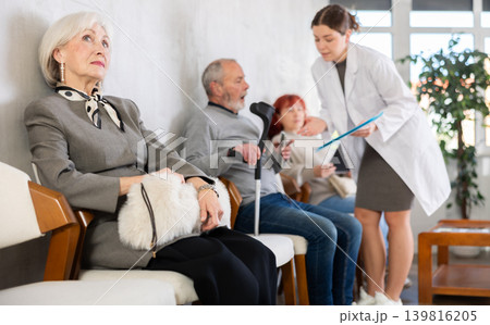 Group of elderly people sitting in chairs in queue to see doctor, nurse talking with patients in background in lobby medical facility 139816205