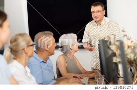 Positive teacher conducting group lesson of computer skills to seniors Positive teacher conducting group lesson of computer skills to seniors 139816273