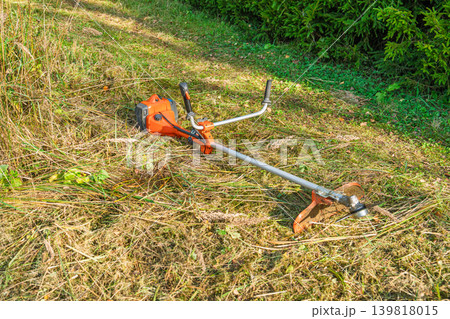 Orange string trimmer resting on freshly mown grass, garden care equipment set out on the lawn Orange string trimmer resting on freshly mown grass, garden care equipment set out on the lawn 139818015