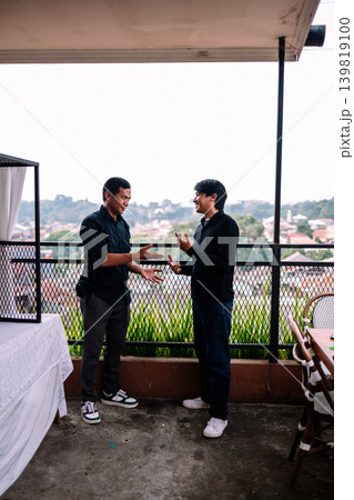 Two Southeast Asian men in black shirts having casual conversation outdoors 139819100