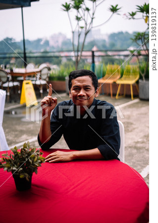 Asian man pointing upward as if having good idea at outdoor table Asian man pointing upward as if having good idea at outdoor table 139819137