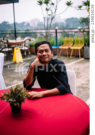 Asian man relaxing with head supported by hand at outdoor table 139819139