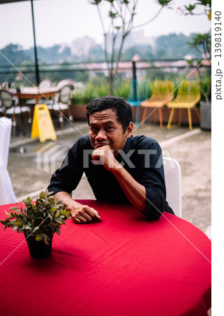Southeast Asian man contemplating with hand on mouth at outdoor table Southeast Asian man contemplating with hand on mouth at outdoor table 139819140