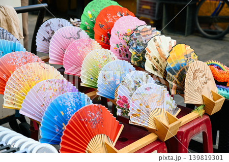 Traditional colorful handmade folding bamboo fans with painted patterns displayed at outdoor souvenir street market booth in Vietnam Traditional colorful handmade folding bamboo fans with painted patterns displayed at outdoor souvenir street market booth in Vietnam 139819301
