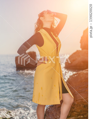 Woman ocean sunset standing on rocky shore in yellow vest during golden hour Woman ocean sunset standing on rocky shore in yellow vest during golden hour 139820020