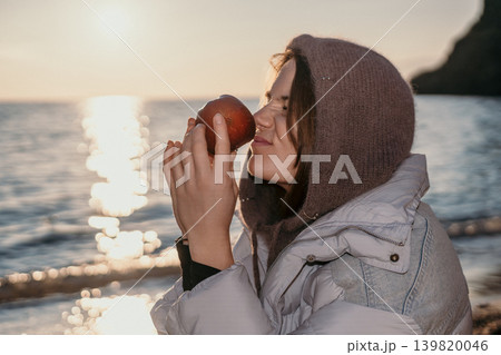 Woman apple beach smelling fresh fruit by the sea during a golden sunset while wearing warm winter clothing and a hood Woman apple beach smelling fresh fruit by the sea during a golden sunset while wearing warm winter clothing and a hood 139820046