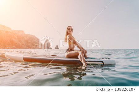 Woman Paddleboard Ocean - Young woman in a bikini sits on a paddleboard in the ocean on a sunny day. 139820107