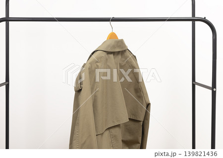Beige women trench coat hanging on rack. Stylish outerwear garment placed on hanger. Female spring wardrobe clothing for modern fashion outfit.. Beige women trench coat hanging on rack. Stylish outerwear garment placed on hanger. Female spring wardrobe clothing for modern fashion outfit.. 139821436