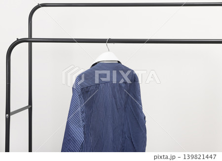 Blue striped shirt hanging on metal rack. Garment suspended from clothing hanger. Male wardrobe minimal fashion clothing concept.. 139821447