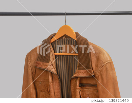 Brown leather jacket hanging on hanger below rail. Outerwear garment displayed on clothing rack. Menswear wardrobe option for transitional weather outfits.. Brown leather jacket hanging on hanger below rail. Outerwear garment displayed on clothing rack. Menswear wardrobe option for transitional weather outfits.. 139821449