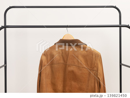Brown leather jacket hanging on metal rack. Outerwear garment placed on wooden hanger. Menswear wardrobe choice for autumn spring season.. Brown leather jacket hanging on metal rack. Outerwear garment placed on wooden hanger. Menswear wardrobe choice for autumn spring season.. 139821450