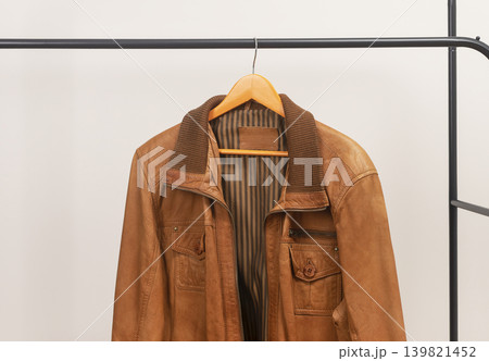 Brown leather jacket hanging on rack with zipper. Outerwear garment placed on wooden hanger. Menswear wardrobe clothing for autumn spring season.. 139821452