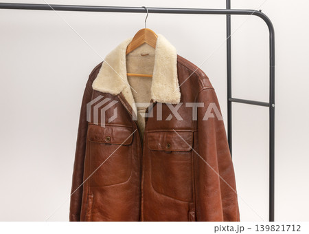 Brown leather jacket hangs on wooden hanger against white background. Shearling collar outerwear rests on black metal rack creating wardrobe display. Minimalist retail fashion concept for Brown leather jacket hangs on wooden hanger against white background. Shearling collar outerwear rests on black metal rack creating wardrobe display. Minimalist retail fashion concept for 139821712