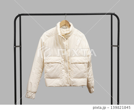 Beige puffer jacket hangs on wooden hanger within black metal rack against grey background. Quilted garment forms structured wardrobe display on horizontal rail. Minimalist retail fashion 139821845