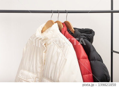 Beige and red puffer jackets hang on metal rack against white background. Quilted outerwear rests on wooden hangers creating wardrobe display. Minimalist retail fashion concept for boutique Beige and red puffer jackets hang on metal rack against white background. Quilted outerwear rests on wooden hangers creating wardrobe display. Minimalist retail fashion concept for boutique 139821850