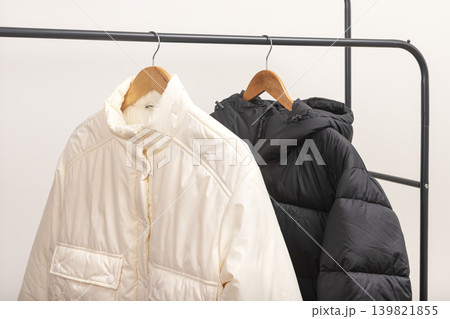 Beige and black puffer jackets hang on metal rack against white background. Quilted outerwear rests on wooden hangers forming retail wardrobe display. Minimalist fashion concept for boutique Beige and black puffer jackets hang on metal rack against white background. Quilted outerwear rests on wooden hangers forming retail wardrobe display. Minimalist fashion concept for boutique 139821855