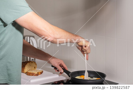 Mature man stirs eggs in frying pan while cooking breakfast at stove. Adult male prepares meal during simple domestic kitchen routine. Everyday home cooking activity in modern interior.. 139822587