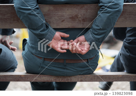 A close-up, back view of a person wearing a grey work uniform or jacket, sitting on a wooden bench. Candid shot of a person observing an event with hands folded behind. A close-up, back view of a person wearing a grey work uniform or jacket, sitting on a wooden bench. Candid shot of a person observing an event with hands folded behind. 139823098