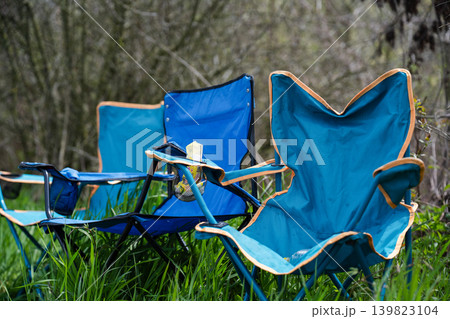 A row of blue foldable camping chairs with cup holders set up on a lush green lawn.  Evoking feelings of relaxation, picnic, or outdoor event waiting to happen. 139823104