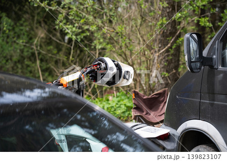A white professional motocross helmet rests on the handlebar of a dirt bike, parked between two vehicles in a forest setting. The scene captures the essence of a weekend adventure or a racing event 139823107