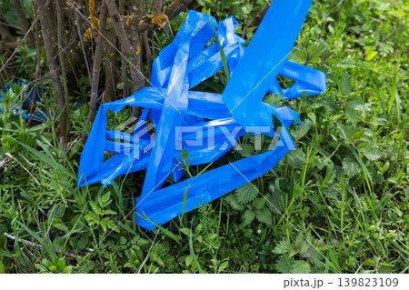 Blue plastic tape discarded in green grass and bushes. Plastic waste littering the natural environment. Blue plastic tape discarded in green grass and bushes. Plastic waste littering the natural environment. 139823109