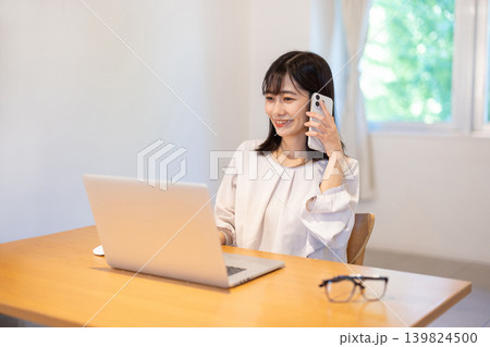 パソコンで作業しながらスマートフォンで通話する日本人の若い女性 139824500