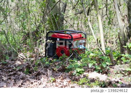 Portable red gasoline generator standing on the ground in a green forest.  Portable power station for camping and remote work.  Off-grid energy source concept 139824612