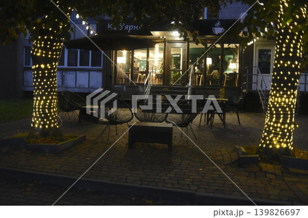 Tables and chairs sit outside a cafe at night. Lights decorate the trees Tables and chairs sit outside a cafe at night. Lights decorate the trees 139826697