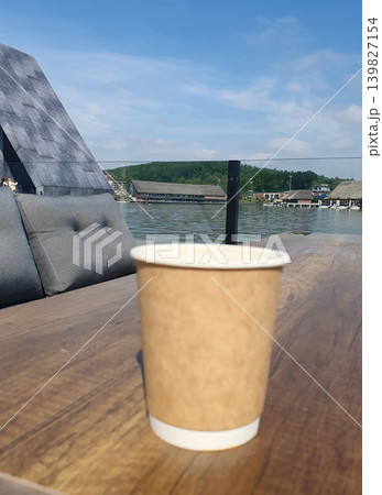 Paper cup of coffee on wooden table of terrace by lake. Cozy seating area with pillows Paper cup of coffee on wooden table of terrace by lake. Cozy seating area with pillows 139827154