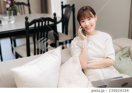 Happy young asian woman sitting on comfortable sofa talking on smartphone using laptop computer smiling bright modern living room home office communication freelance lifestyle telecommute 139829184
