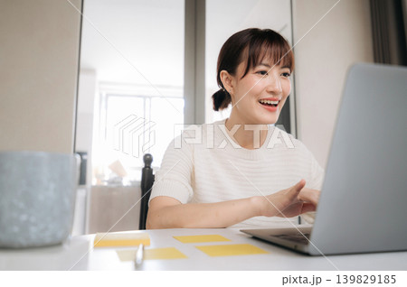 Happy young asian woman working on laptop computer at home office desk showing joyful emotional expression during online video meeting using modern internet communication technology Happy young asian woman working on laptop computer at home office desk showing joyful emotional expression during online video meeting using modern internet communication technology 139829185