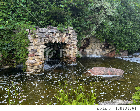 Waterfall cascading over stone structure surrounded by lush greenery in serene garden setting Waterfall cascading over stone structure surrounded by lush greenery in serene garden setting 139831361
