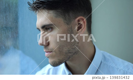 Portrait of sad young entrepreneur sitting near window in modern hotel room or office. Unhappy man with serious expression looking away showing emotional stress, fatigue and professional burnout Portrait of sad young entrepreneur sitting near window in modern hotel room or office. Unhappy man with serious expression looking away showing emotional stress, fatigue and professional burnout 139831432