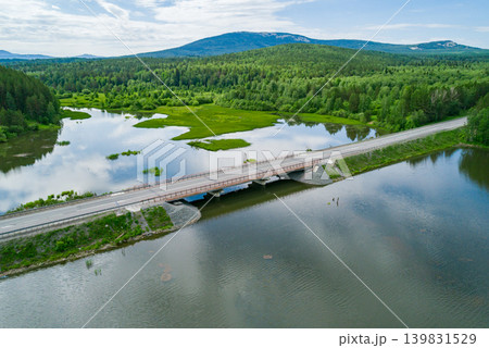 Concrete bridge extends over a wide river, connecting forested shores and leading to distant green mountains 139831529