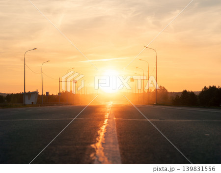 An empty road leads toward the horizon at golden hour, with the sun low in the sky creating a bright glare An empty road leads toward the horizon at golden hour, with the sun low in the sky creating a bright glare 139831556