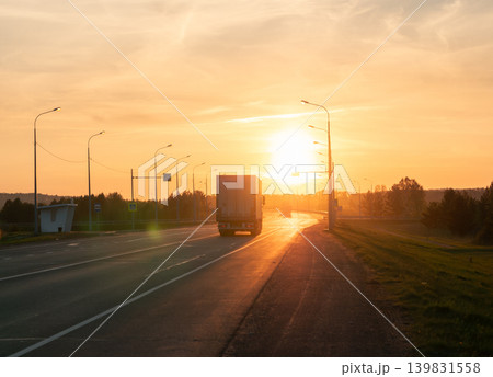 A large semitruck drives along a multi-lane highway against bright sunlight at sunset A large semitruck drives along a multi-lane highway against bright sunlight at sunset 139831558