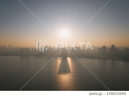 Aerial views of a Chinese cityscape at sunrise show buildings rising above a river. Early morning sun rays shine through the city haze. 139831648