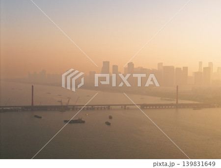 Morning haze blankets a sprawling urban landscape featuring a long bridge across a wide river with distant skyscrapers Morning haze blankets a sprawling urban landscape featuring a long bridge across a wide river with distant skyscrapers 139831649
