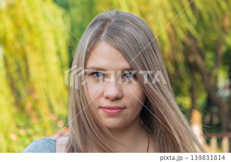 A young woman with straight light blonde hair and blue eyes looks directly forward. She is outdoors against a softly blurred background of green and yellow foliage. 139831814