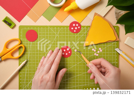 Top view of hands using tweezers to assemble a paper pizza pepperoni on a green cutting mat. Creative DIY workspace with colorful paper, scissors, glue, and stationery for kids art projects. 139832171