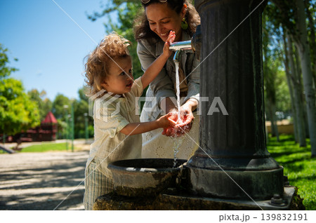 Mother helps her young son wash hands under a park fountain, sharing a joyful moment on a sunny day at the playground with family bonding and summer fun. Mother helps her young son wash hands under a park fountain, sharing a joyful moment on a sunny day at the playground with family bonding and summer fun. 139832191
