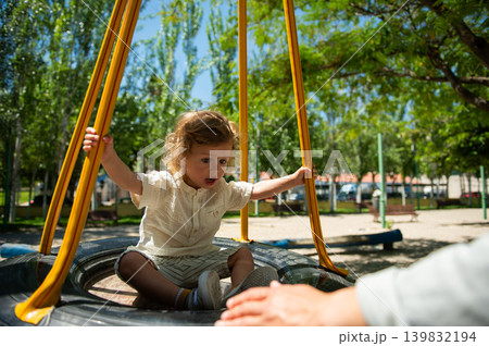 Toddler on a tire swing reaches for a parent's hand in a sunlit playground, capturing playful, curious and joyful family moments during a warm summer day outdoors. Toddler on a tire swing reaches for a parent's hand in a sunlit playground, capturing playful, curious and joyful family moments during a warm summer day outdoors. 139832194
