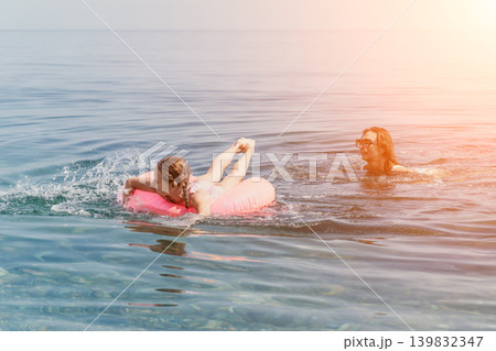Family beach swimming mother and young daughter enjoy a sunny day playing with a pink inflatable ring in the calm blue sea water Family beach swimming mother and young daughter enjoy a sunny day playing with a pink inflatable ring in the calm blue sea water 139832347